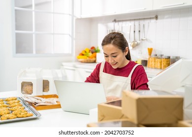 Asian Woman Bakery Shop Owner Using Laptop Computer With Internet Presentation Dessert Bakery On Social Media In The Kitchen. Small Business Entrepreneur And Online Marketing Food Delivery Concept