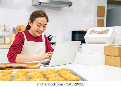 Asian Woman Bakery Shop Owner Using Laptop Computer With Internet Presentation Dessert Bakery On Social Media In The Kitchen. Small Business Entrepreneur And Online Marketing Food Delivery Concept