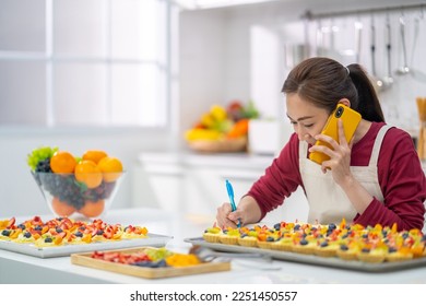 Asian woman bakery shop owner talking on mobile phone to customer and preparing customer order selling online delivery in the  kitchen. Small business food and drink occupation entrepreneur concept. - Powered by Shutterstock