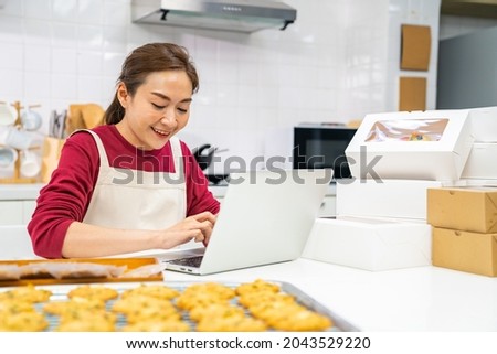Asian woman bakery shop owner using laptop computer with internet presentation dessert bakery on social media in the kitchen. Small business entrepreneur and online marketing food delivery concept