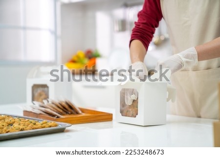 Asian woman bakery shop owner preparing customer order cookie in delivery box on kitchen counter. Woman bakery chef making bakery in the kitchen. Small business entrepreneur and food delivery concept.