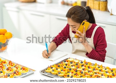 Asian woman bakery shop owner talking on mobile phone to customer and preparing customer order selling online delivery in the  kitchen. Small business food and drink occupation entrepreneur concept.