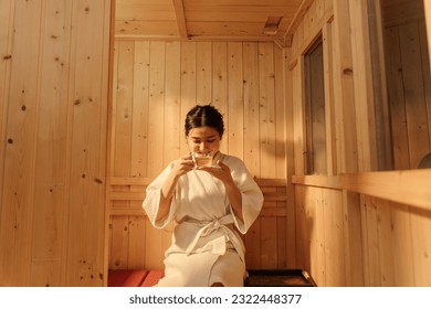 Asian woman in bathrobe drinking hot tea while relaxing in the sauna. Female healthcare and spa treatment concept. - Powered by Shutterstock