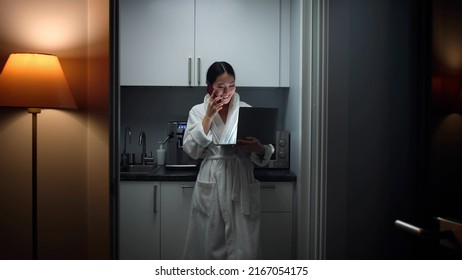 Asian Woman In Bathrobe Talk On Smartphone And Work On Computer Standing In Kitchen Late In Evening