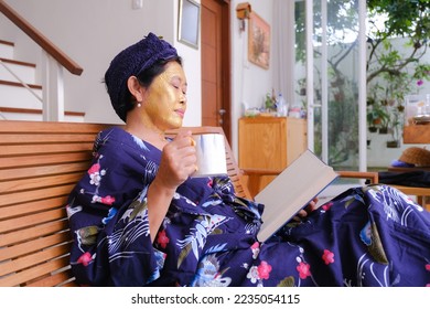 An Asian woman with a beauty mask on her face is holding a cup while reading a book. - Shutterstock ID 2235054115