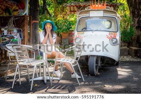 Asian woman in big hat sitting on chair with tuk tuk Thailand in conner of open air restaurant