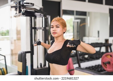 An Asian Woman In A Black Crop Top Looks At The Camera While Doing Dynamic Chest Stretch Before A Workout Session At The Gym.