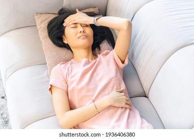 Asian Woman With Black Hair Having A Headache Lying On The Grey Sofa, Sick At Home, With Stomach Pain, Sensitive To Weather, Unwell, During Menstruation, Hold On To Your Stomach, Your Head, Resting