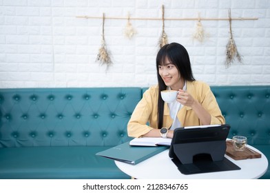Asian Woman with black hair working comfortably with a tablet  drinking hot coffee.A young Asian girl sits at work relaxing with her favorite cup of coffee.Business woman enjoying to drink beverage.