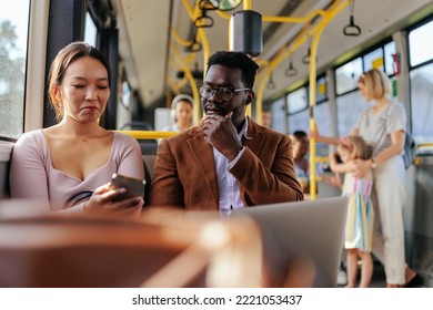 An Asian Woman And A Black Man Are Riding On A City Bus And Looking At A Smartphone