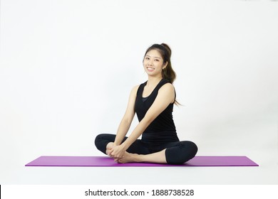 Asian Woman In Black Sportswear Sitting On Purple Yoga Mat Looking Camera And Pose On The White Background.