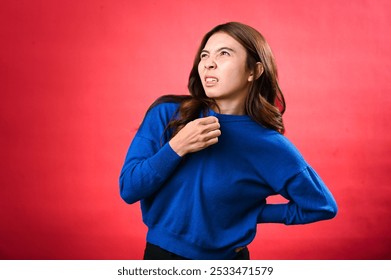 An Asian woman in a blue sweater appears uncomfortable, as she pulls at her collar and touches her back, expressing heat or discomfort. The background is solid red - Powered by Shutterstock