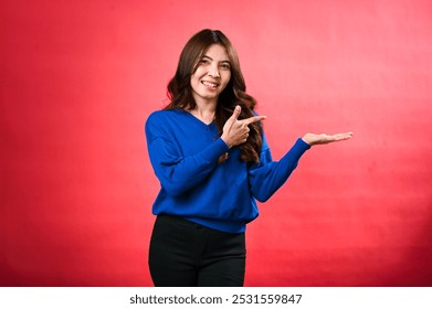 An Asian woman in a blue sweater presents with one hand outstretched and the other pointing towards it. She smiles confidently, standing against a bright red background in a casual, friendly pose - Powered by Shutterstock