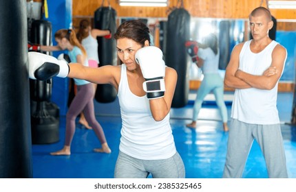 Asian woman in boxing gloves exercising jabs with punchbag during her boxing training. Her trainer standing behind and observing. - Powered by Shutterstock