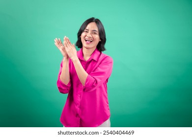 An Asian woman in a bright pink shirt stands against a green background, laughing and clapping her hands, showing joy and excitement. Her pose and smile radiate positivity and friendliness. - Powered by Shutterstock