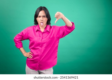 An Asian woman in a bright pink shirt poses against a green background, making a playful face by sticking out her tongue and giving a thumbs-down gesture. Her expression is humorous