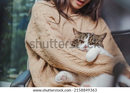 Asian woman in brown sweater holding adorable white with brown and black strip cat in her arm, cat in human hug embrase human arm and looking at camera.