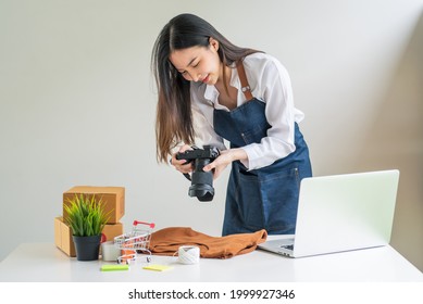 Asian woman business owner holding a camera to take pictures of products to sell online a laptop and parcel box at home. - Powered by Shutterstock