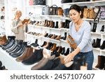 Asian woman buying new footwear in shoe store. European granny choosing shoes in background.