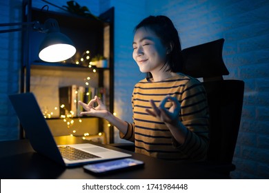 Asian woman calms down, breathes deeply with meditating holding her hands in yoga gesture after working on a laptop at the night at home. WFH. Work from home avoid COVID 19 concept.