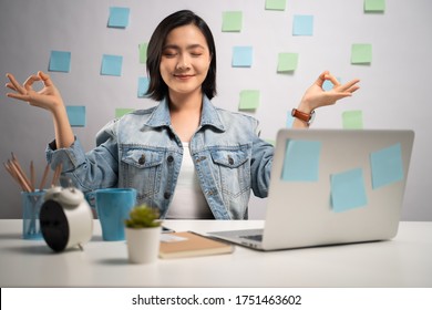 Asian woman calms down, breathes deeply with meditating holding her hands in yoga gesture after working on a laptop at home office.
