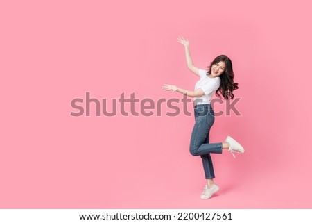 An asian woman in a casual outfit hopping off with her arm presenting, a cheerful expression on her face as she is excited to present something in isolated pink background.