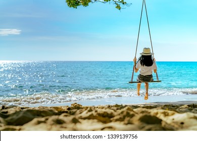 Asian woman in casual style wear hat swing the swings at sand beach  and looking beautiful tropical paradise sea and sky on sunset. Summer vacation. Summer vibes. Enjoying and relaxing girl on holiday