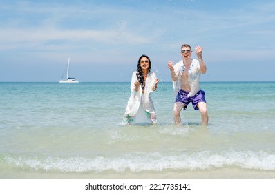 Asian Woman With Caucasian Man Splash Water To Camera And Look Fun Together With Some Boat On Background On The Sea With Clear Blue Sky.