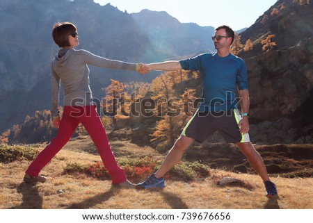 Asian woman and caucasian man in sport mountaineering trekking outfit & sunglasses holding hands do gymnastic stretching sport together in mountain autumn landscape sunny day  Bardonecchia Turin Italy