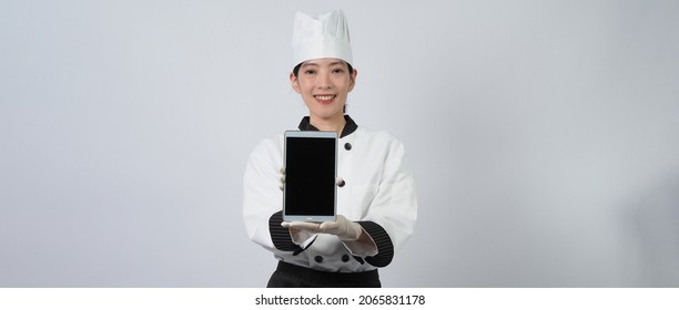 Asian Woman Chef Holding Smartphone Or Digital Tablet And Received Order From Online Shop Or Merchant Application. She Smiling In Chef Uniform And Standing In White Background. Online Food Merchant. 