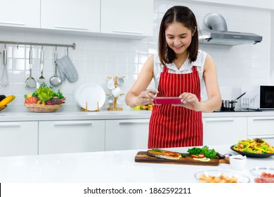 Asian Woman Chef In Red Apron Take A Picture Of Salmon Steak With Smartphone. Concept Woman Preparing Meals At Home.