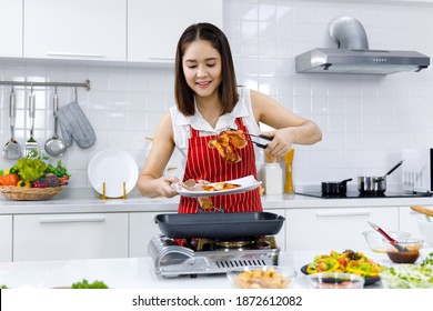 Asian Woman Chef In Red Apron Cooking Steak In Kitchen At Home. Concept Woman Preparing Meals At Home.