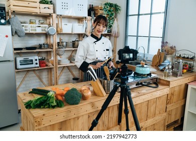 Asian Woman Chef Is Talking To The Digital Camera On Tripod While Filming And Teaching Cooking Online In A Bright Rustic Kitchen At Home