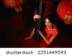 asian woman in Chinese costume playing chinese harp or Guzheng with background of red umbrella and latern to celebrate Chinese new year, translation: wish you prosperity and wealth