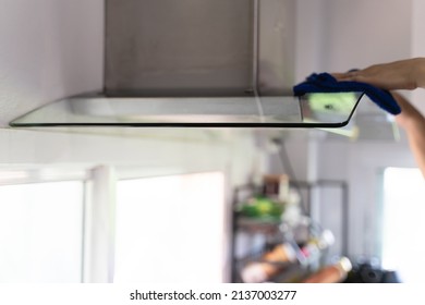 Asian Woman Cleaning Dirty Dust With A Blue Microfiber Cloth On The Kitchen Hood Fan Glass. A Common Household Chores For Housewife.