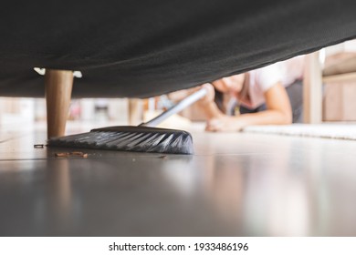 Asian woman cleaning and sweeping dust the floor under the sofa with a broom in the living room. Woman doing chores at home. Housekeeping concept.