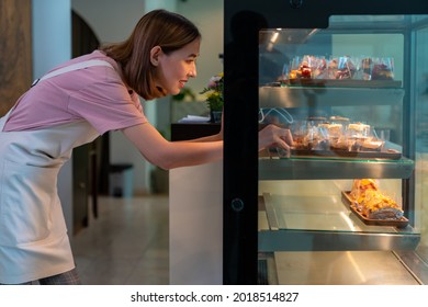 Asian woman coffee shop employee placing pastry and cake in bakery refrigerator showcase at cafe. Female waitress preparing restaurant for service to customer. Small business and part time job concept