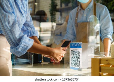 Asian Woman Customer Using Mobile Scan QR Or NFC Payment On Smart Phone Paying For Her Coffee Drink In Coffee Shop And Plexi Screen And Barista Wearing Mask In Background
