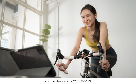 Asian Woman Cyclist Exercise In Home Gym And Watching Tablet During Work Out