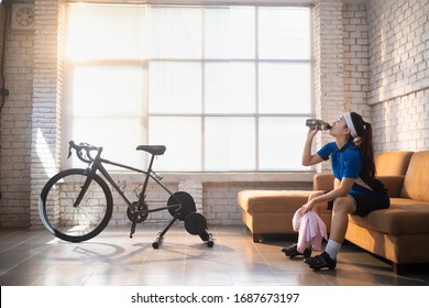 Asian woman cyclist. She is exercising in the house. By cycling on the trainer And play online bike games She breaks to drink water