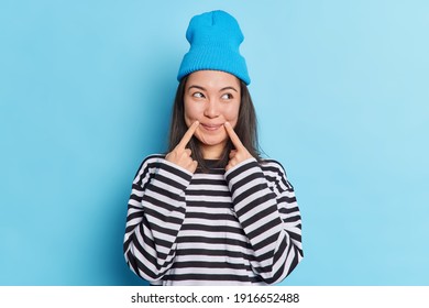 Asian Woman With Dark Hair Keeps Fingers Near Corner Of Lips Has Satisfied Expression Looks Aside Dressed In Long Sleeved Striped Jumper And Hat Has Fake Smile On Face Isolated Over Blue Background