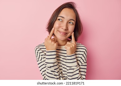 Asian Woman With Dark Hair Keeps Fingers Near Corner Of Lips Has Satisfied Expression Looks Aside Dressed In Long Sleeved Striped Jumper And Hat Has Fake Smile On Face Isolated Over Blue Background