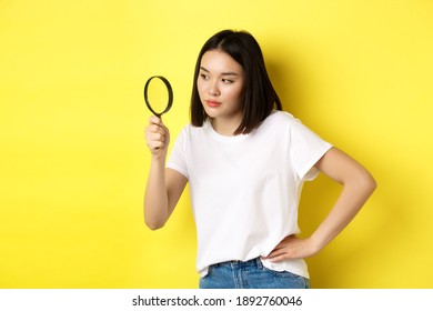 Asian Woman Detective Looking Through Magnifying Glass With Intrigued Look, Found Clues, Standing Over Yellow Background