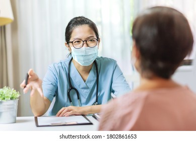 Asian Woman Doctor in blue uniform and protective face mask giving advice to Senior female patient at hospital. Practitioner wear eyeglasses and surgical mask writing information on prescription