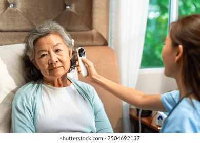 Asian woman doctor examine patient use infrared thermometer measured temperature in senior woman ear. Nurse visit and checking elderly patient health at home. Home medical caregiving service concept. - Powered by Shutterstock