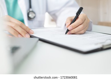 Asian Woman Doctor With Green Cloth And Lab Coat Working And Taking Note Information Of Patient In Checklist Paper On Clipboard In Medical Room Of Hospital. Health Care.