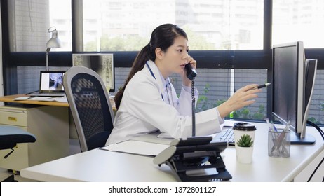 asian woman doctor sitting at desk in front of computer and talking on telephone with a patient telemedicine health hotline consultation. young female medical staff using pen pointing screen discuss