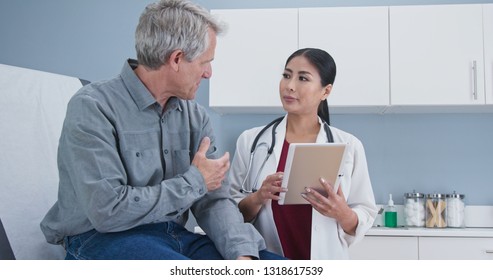 Asian Woman Doctor Talking Senior Caucasian Male Patient Sitting On Exam Table. Medial Professional Using Tablet Computer To Take Medical History From Older Man