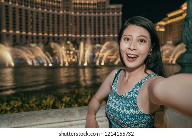 Asian Woman In Dress Taking Selfie Photo With Fountain Water Show In Front The Hotel In Las Vegas. Young Girl Night Out Lifestyle. Fashion Lady Face To Camera Smiling At Night.
