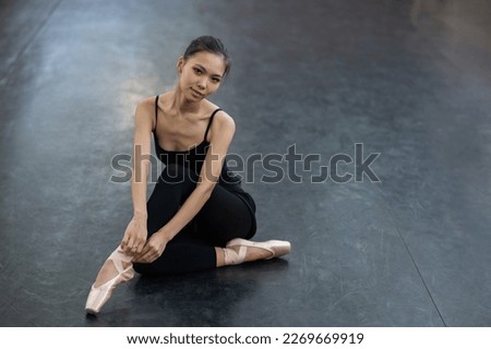An Asian woman dressed in a black bodysuit, leggings and pointe shoes sits on the floor.